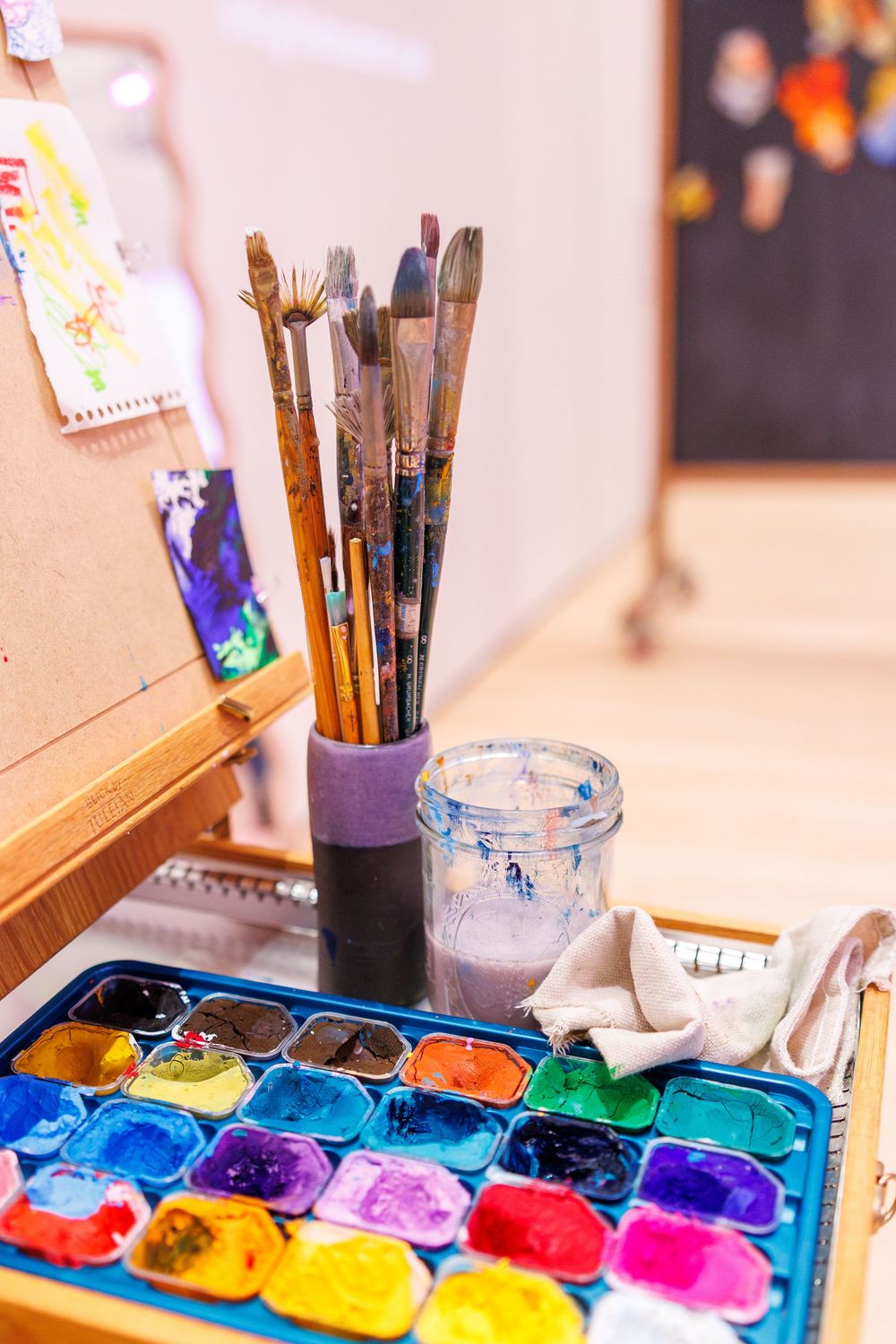 Colorful watercolor paint palette with brushes in a jar and a glass of water on an artist's wooden easel desk.