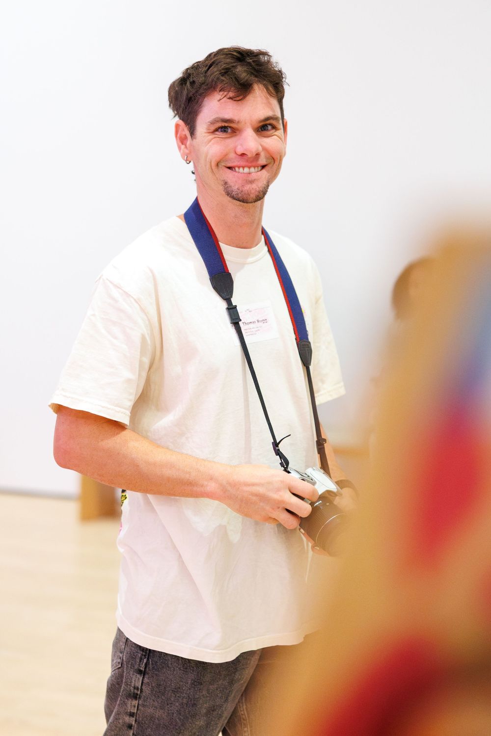 Smiling man in white t-shirt holding a camera with blue strap in a bright gallery space.