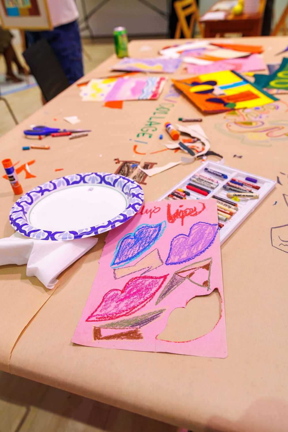 Art supplies and colorful drawings of lips on pink paper spread across a craft table at a children's art workshop.