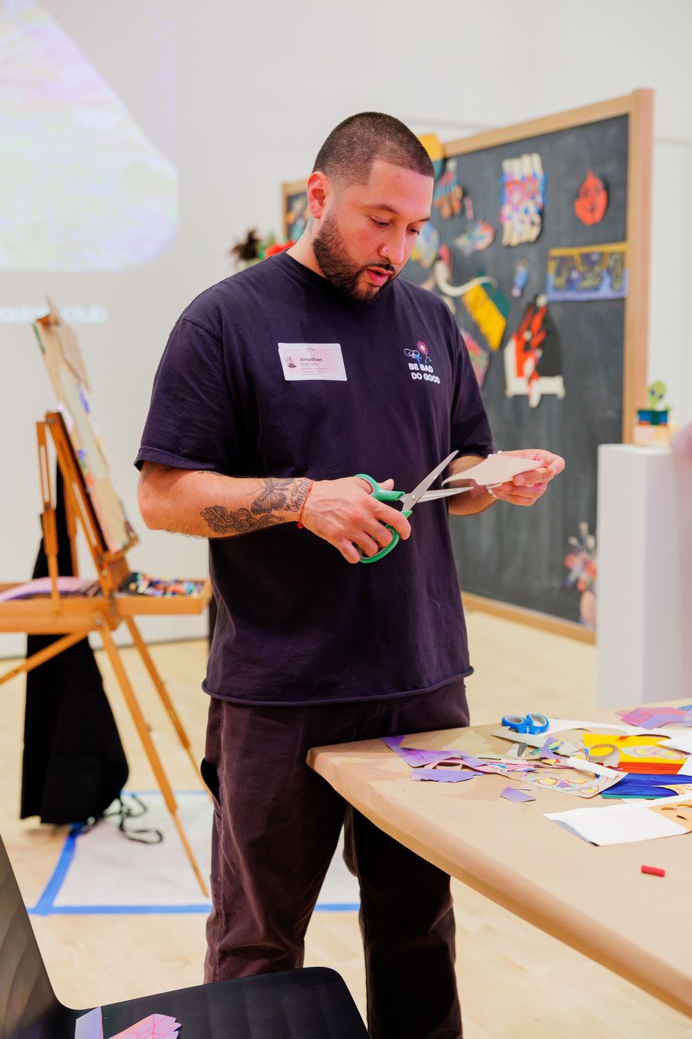 Man with tattoos cutting colorful paper shapes with green scissors at a craft table in an art studio.