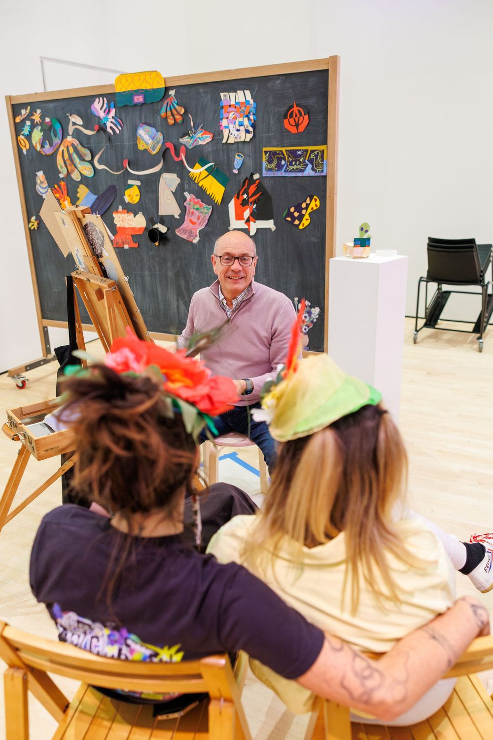Art teacher smiles at two students wearing colorful headbands during a creative workshop in front of a chalkboard.