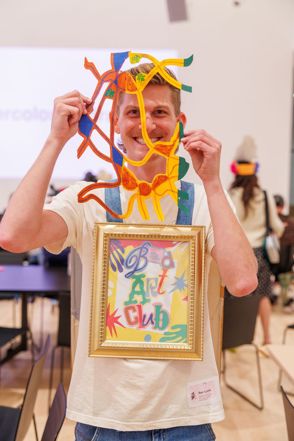 Man smiling and holding colorful pipe cleaner art over his face at a Bad Art Club event.