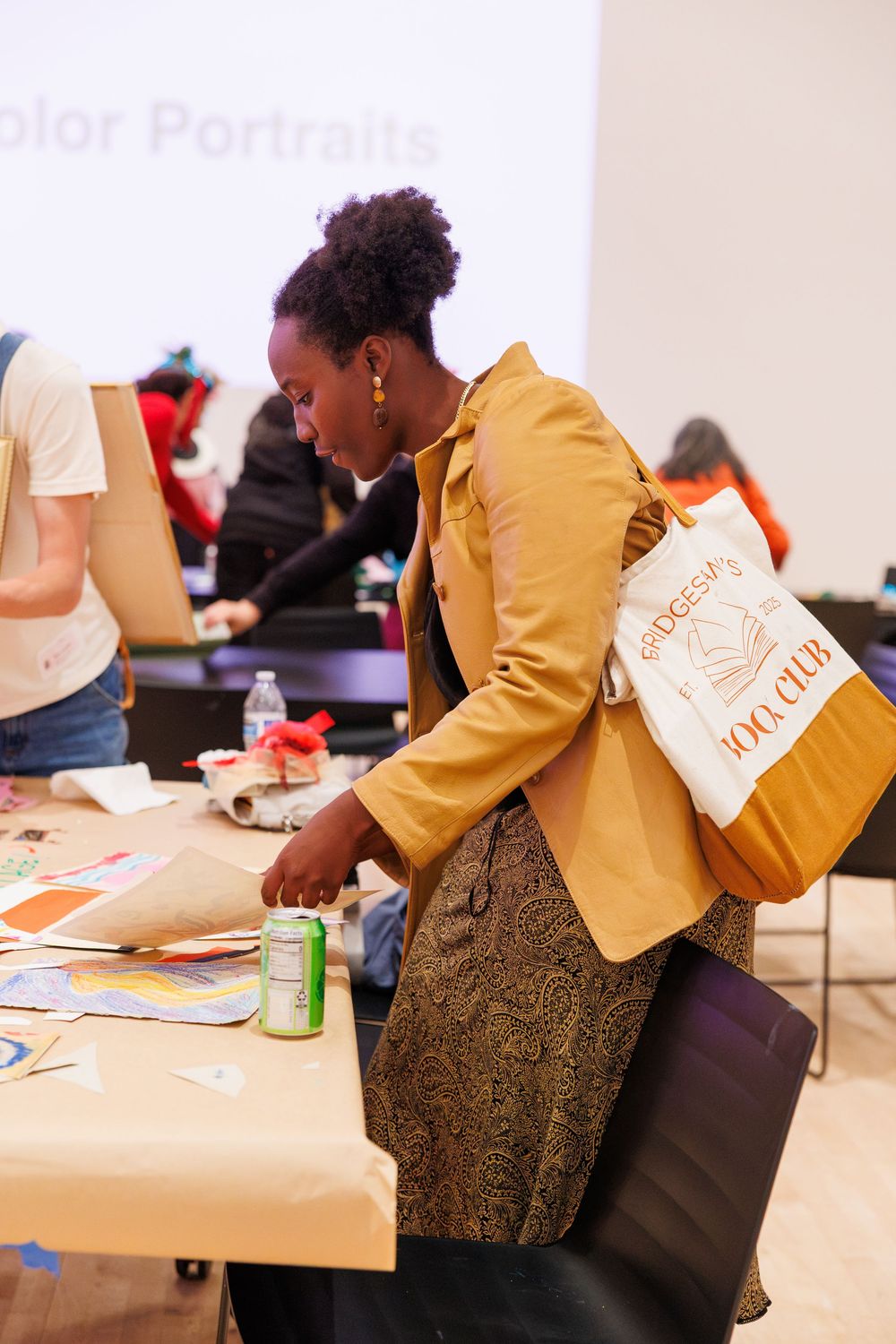 Woman in yellow jacket with tote bag examines art materials at a color portraits workshop table.