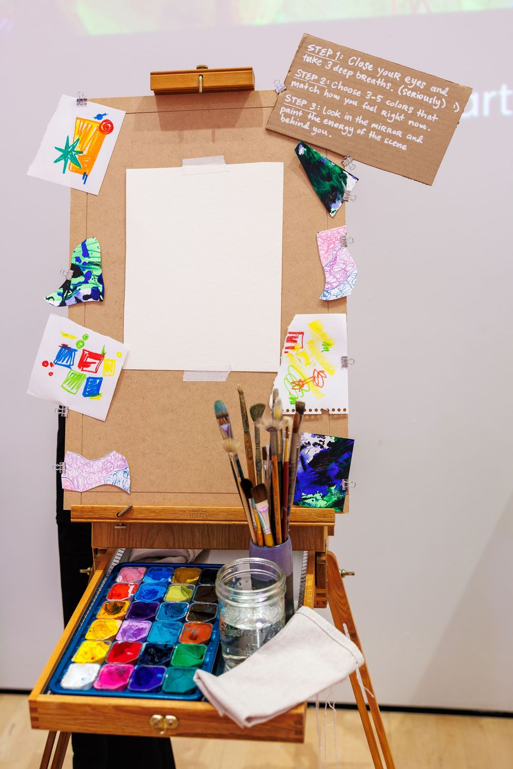 An artist easel with watercolor paints, brushes, and colorful children's artwork displayed on a corkboard with instructions.