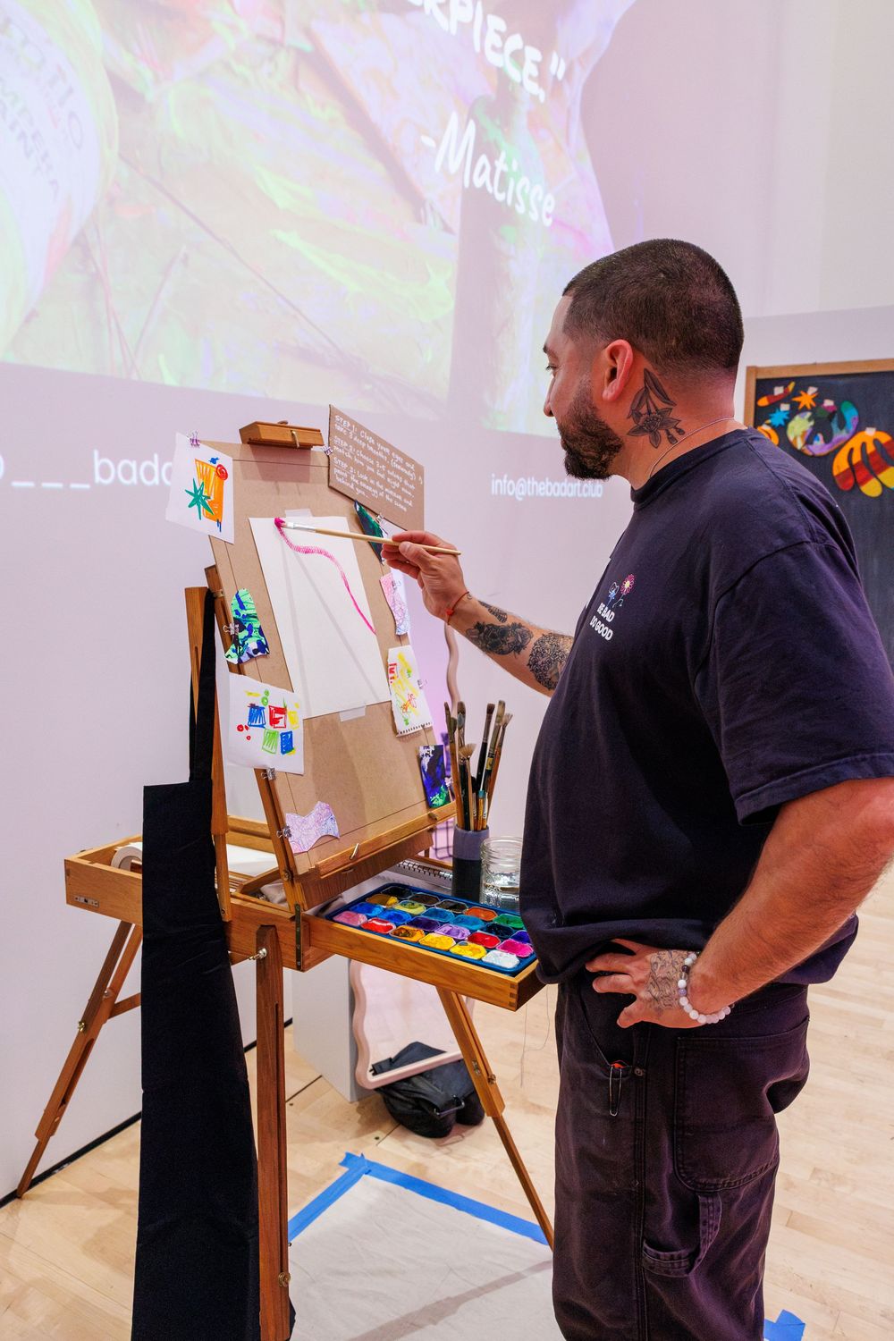 Tattooed male artist painting on easel at art exhibition with colorful palette and Matisse projection backdrop.