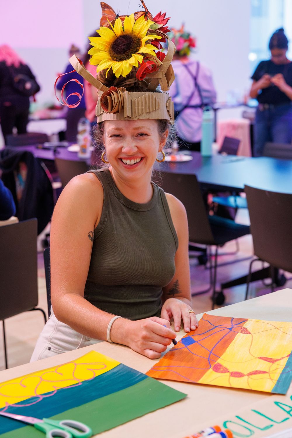 Smiling woman wearing a colorful sunflower crown sits at a table creating vibrant artwork during a creative workshop event.
