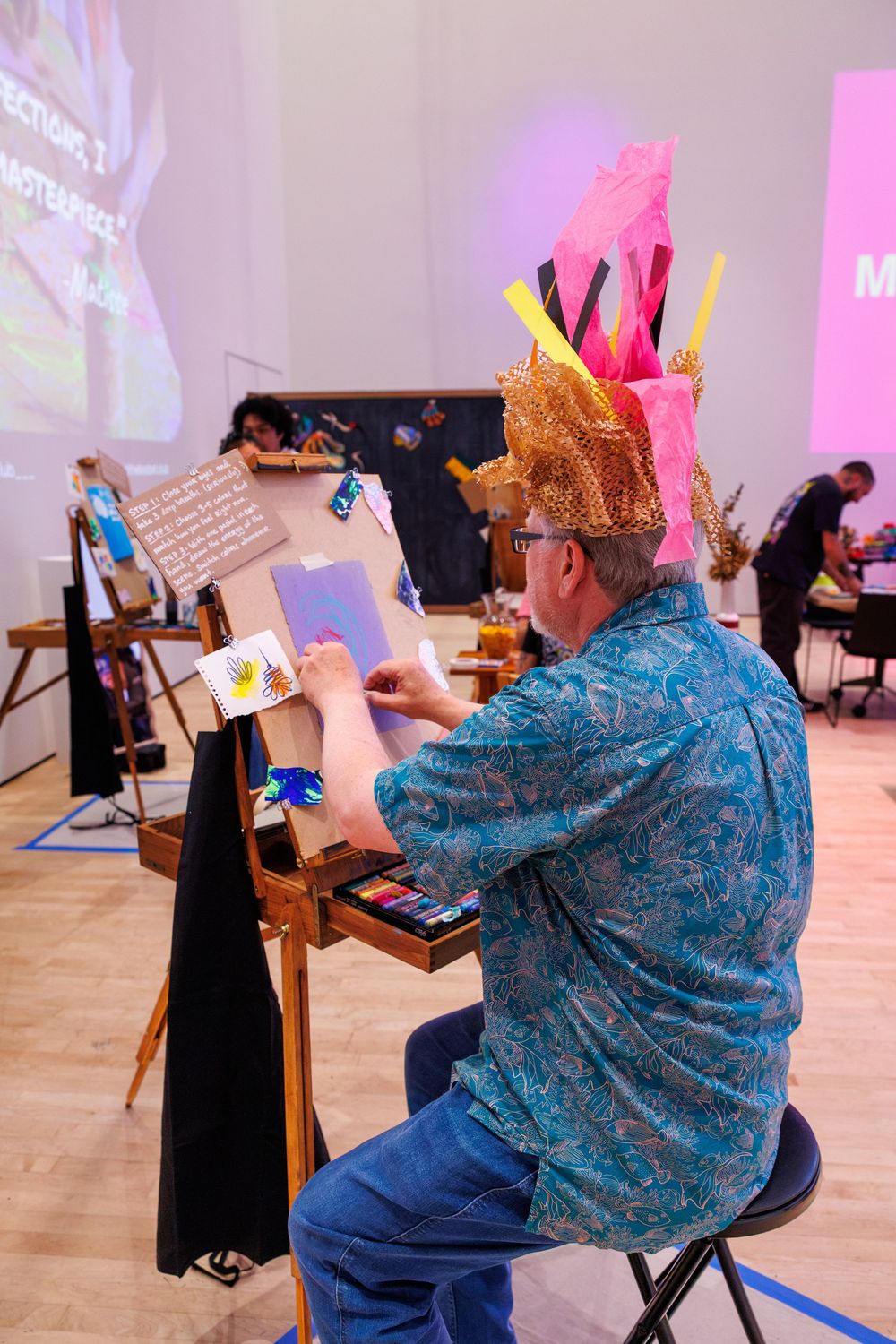 Man wearing a festive pink and gold hat paints at an easel during a lively art competition event.