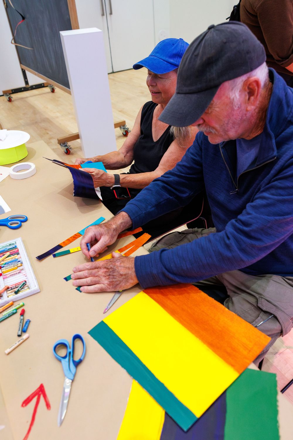 Two seniors sit on floor doing colorful paper craft art project with scissors, markers, and bright colored paper strips.