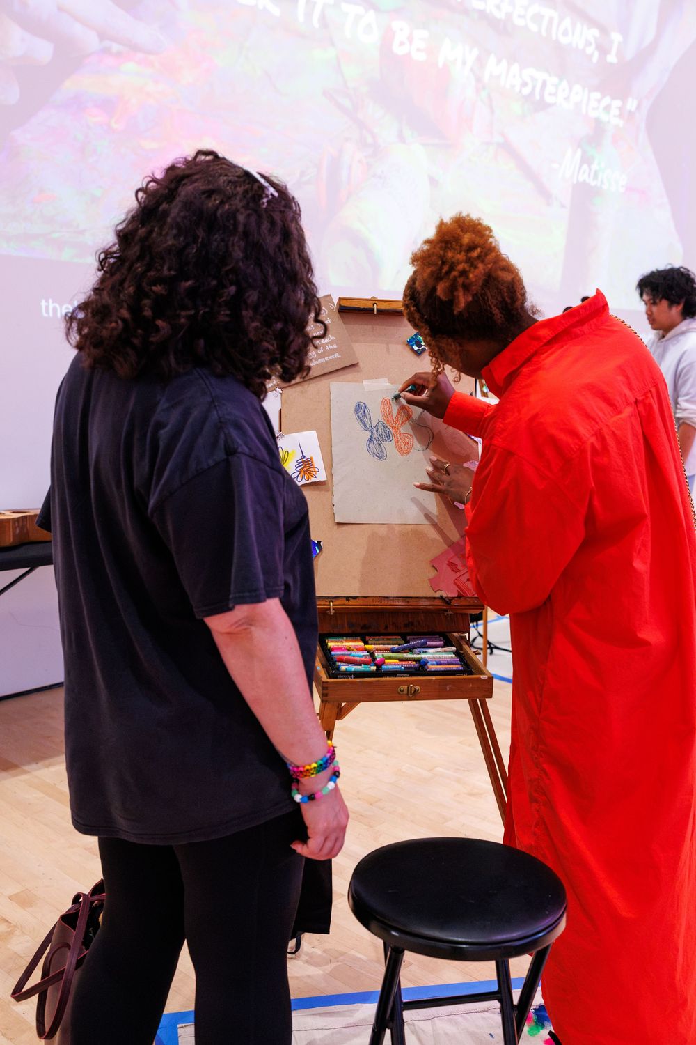 Artist in red jumpsuit demonstrates painting technique to visitor at an art easel during a live gallery event.