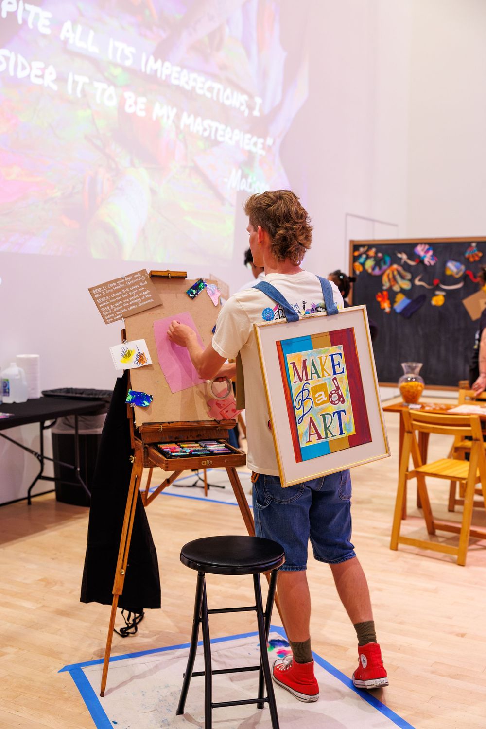 Artist painting at an easel wearing a colorful Make Bad Art framed backpack in a bright creative studio space.