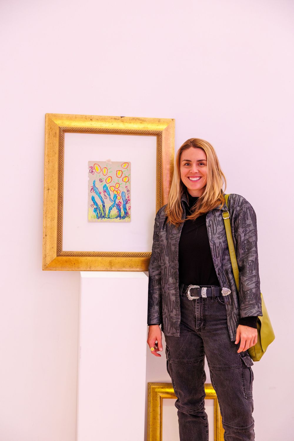 Woman smiling beside a colorful framed artwork on a white pedestal in an art gallery.