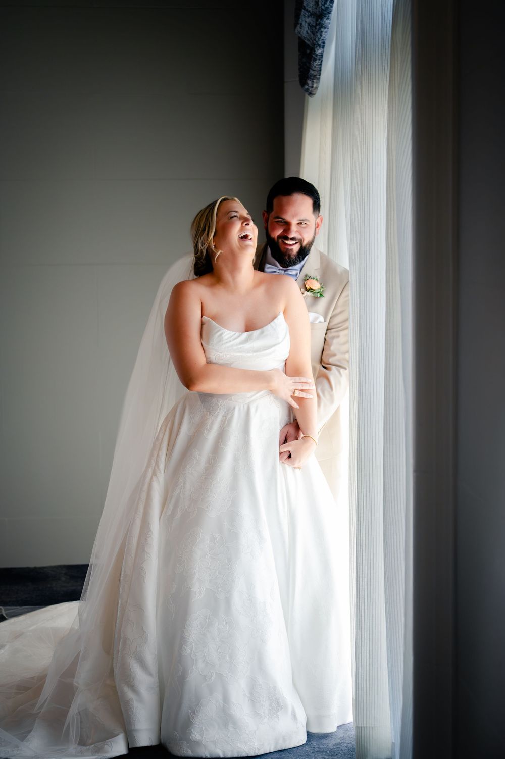 Joyful bride in white strapless gown and veil laughs with groom in tan suit by a sunlit window.