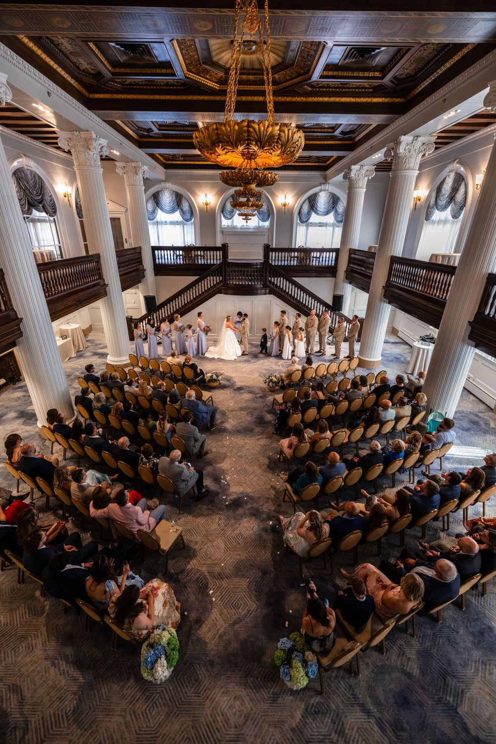 Aerial view of an elegant wedding ceremony in a grand ballroom with white columns, chandelier, and seated guests.