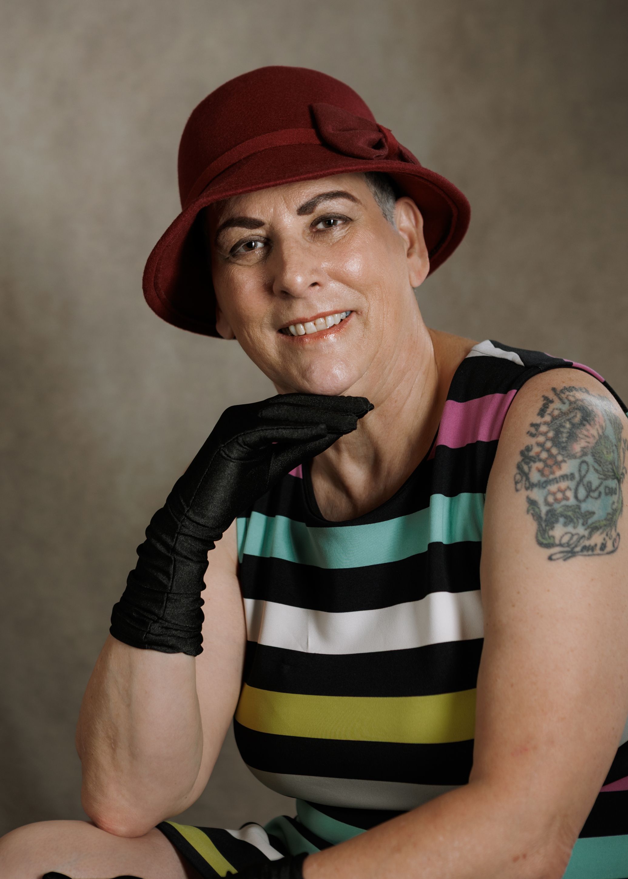 Smiling woman wearing red cloche hat, black gloves, striped dress, and floral tattoo on shoulder.
