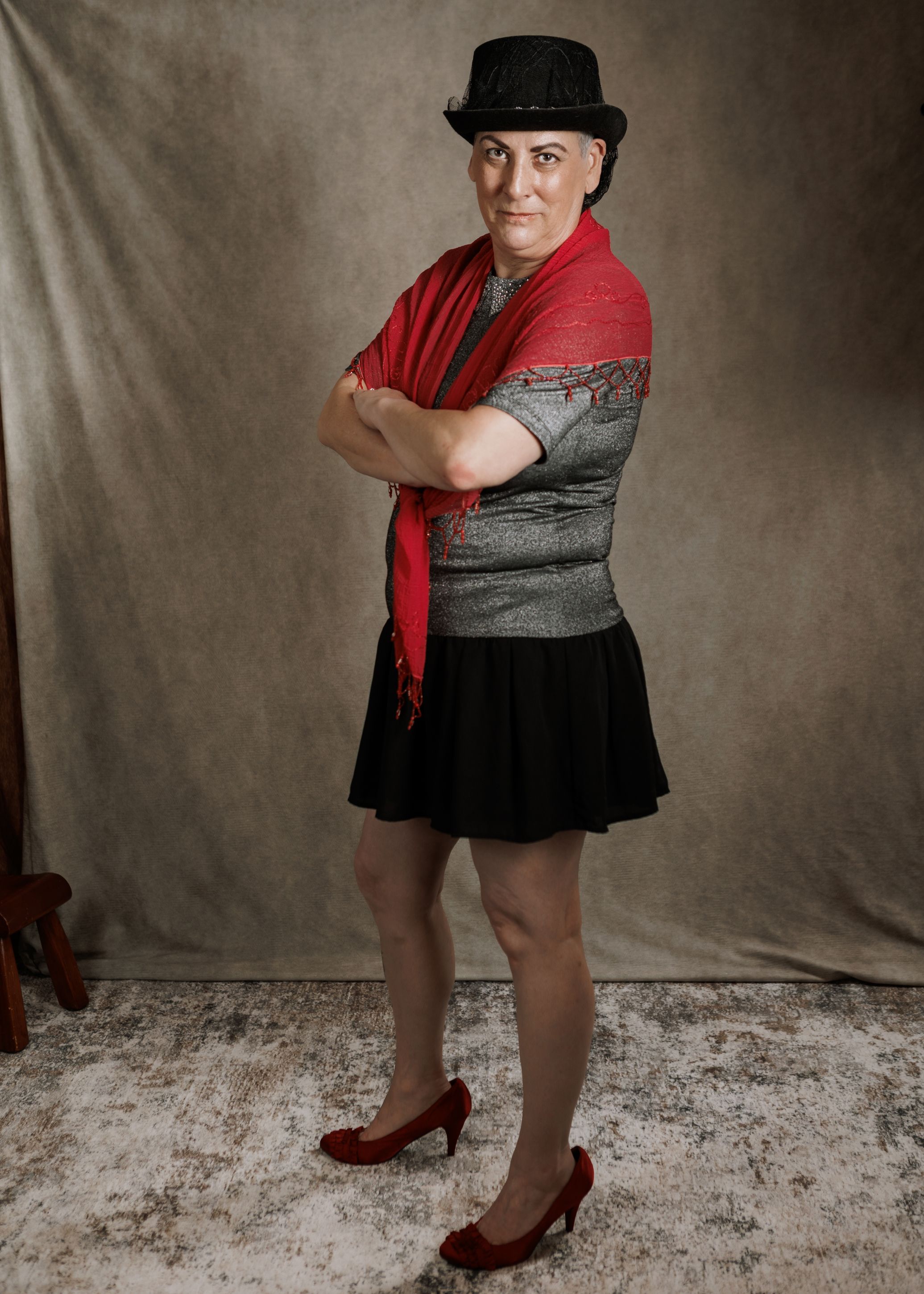 Woman in black skirt, gray top, red scarf, and black top hat poses with arms crossed wearing red heels.