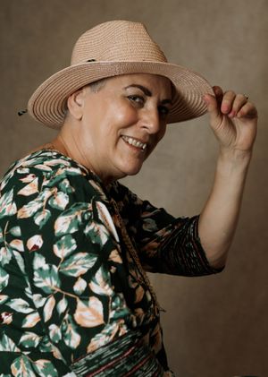 Smiling woman in floral dress tipping a wide-brim straw hat, posing confidently against a neutral background.
