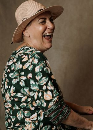Woman in floral green dress and straw hat laughing joyfully, profile view against neutral background.