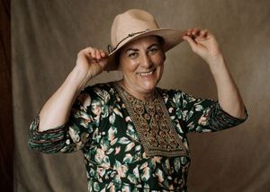 Smiling woman in floral dress adjusting a beige straw hat, posing against a neutral brown backdrop.