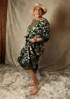 Woman in floral green dress and beige hat poses barefoot against neutral backdrop.