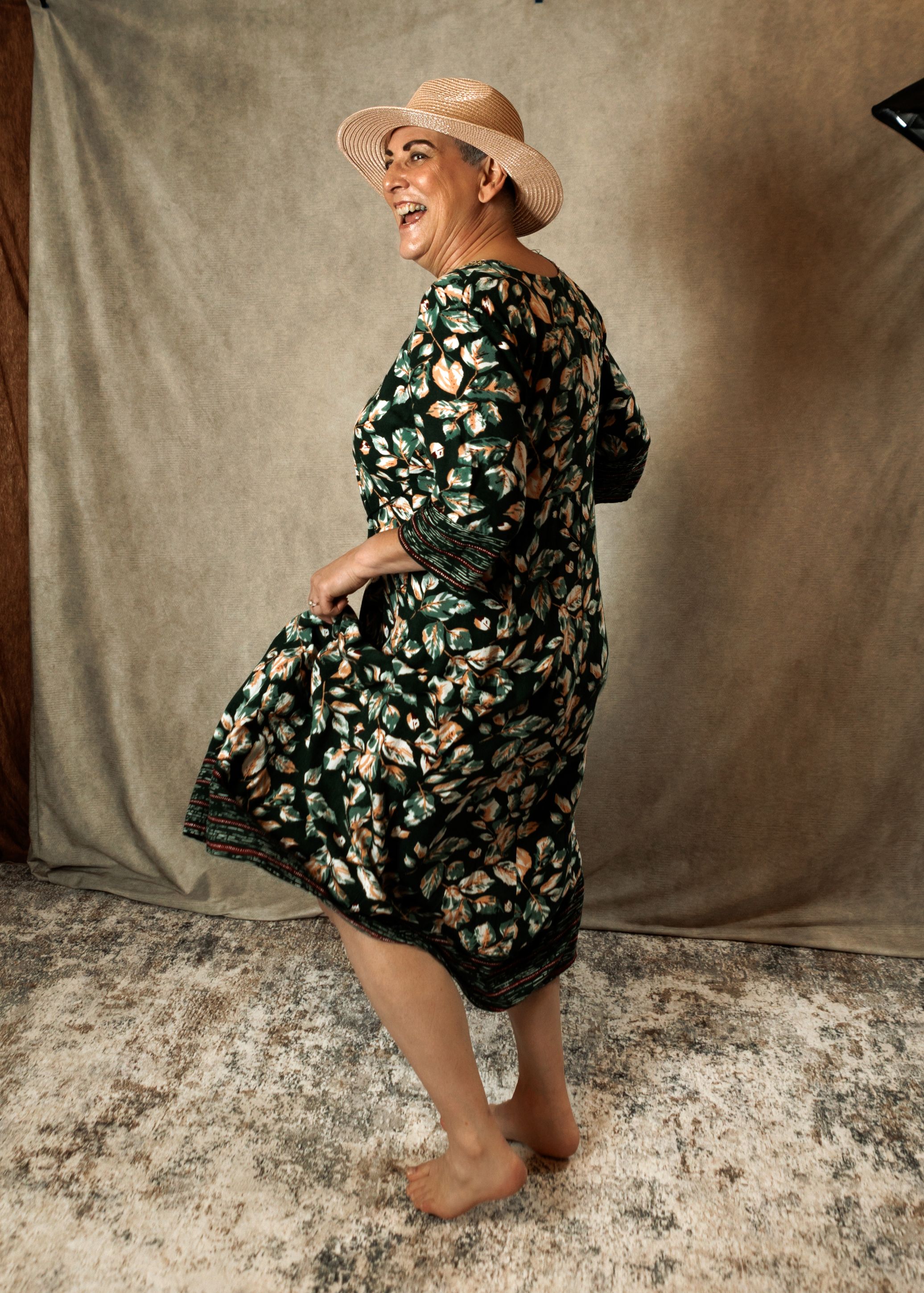Woman in floral dress and tan hat laughing, posing barefoot against neutral studio backdrop as she twirls the skirt of her dress.