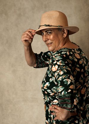 Smiling, cheeky looking woman in floral green dress tipping a straw fedora hat, posing against a neutral studio background.