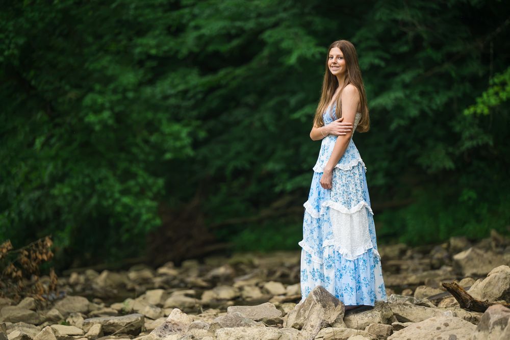 Young woman in blue and white floral maxi dress standing on rocky terrain with lush green trees in background.