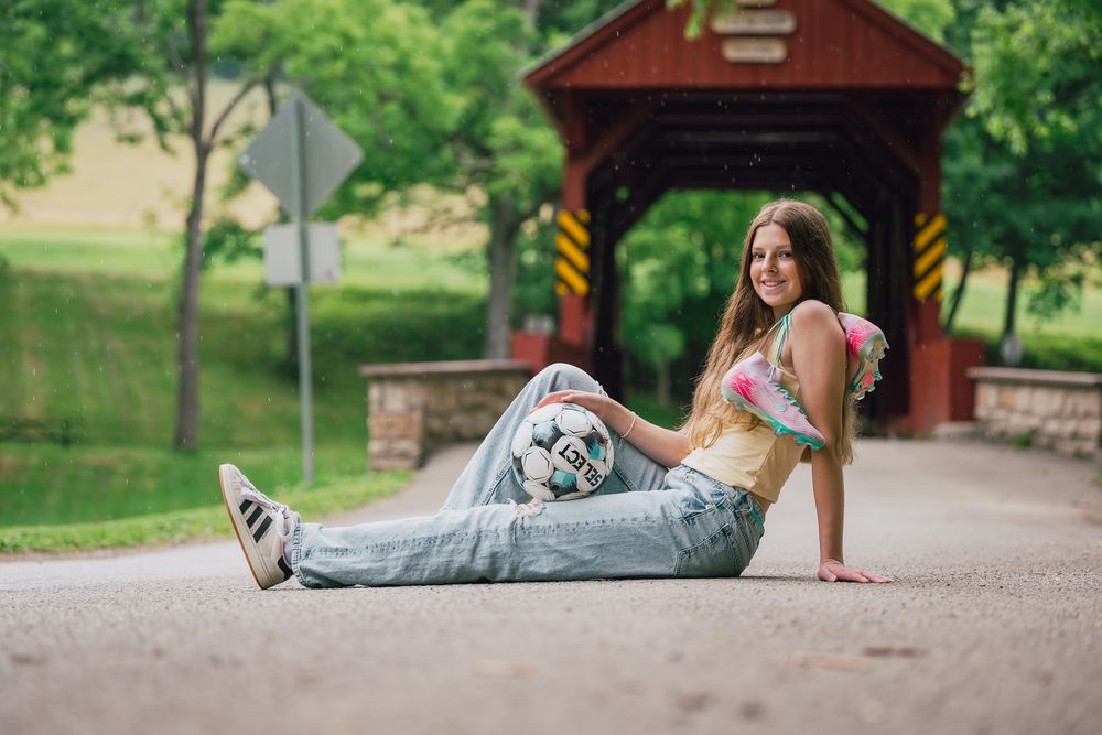 Young woman sitting on a road in jeans and crop top, smiling near a red covered bridge in a green park.