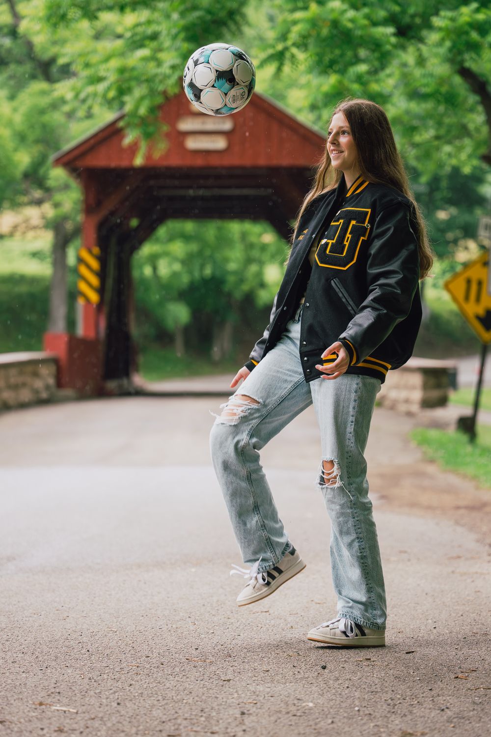 A smiling teen girl in a varsity jacket juggles a soccer ball outdoors near a red covered bridge.