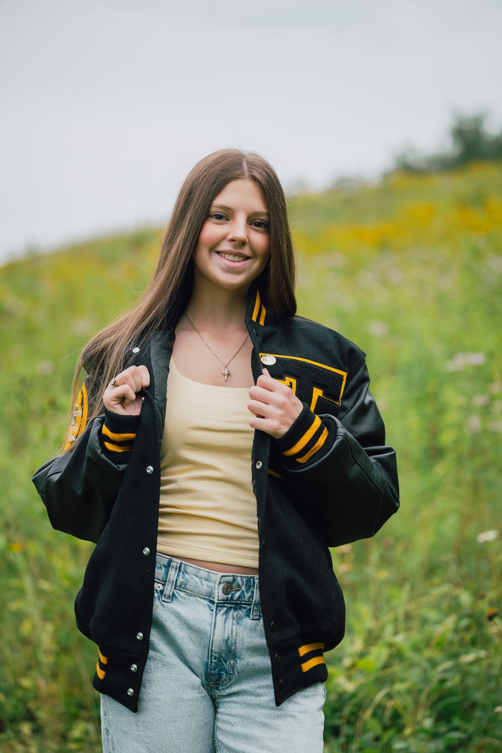 Smiling teenage girl in black and gold varsity jacket and jeans posing in a wildflower field.