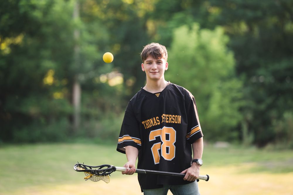 Teen lacrosse player in black Thomas Person #28 jersey tossing a yellow ball while holding a lacrosse stick outdoors.