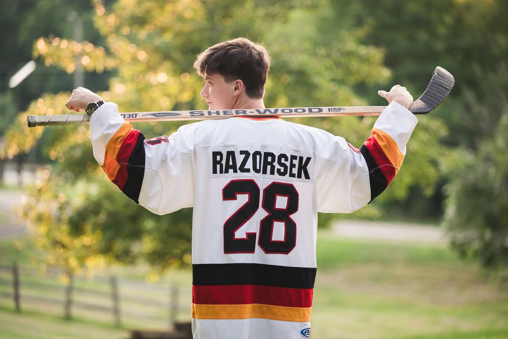 Teen hockey player wearing Razorsek #28 jersey holds stick across shoulders outdoors.