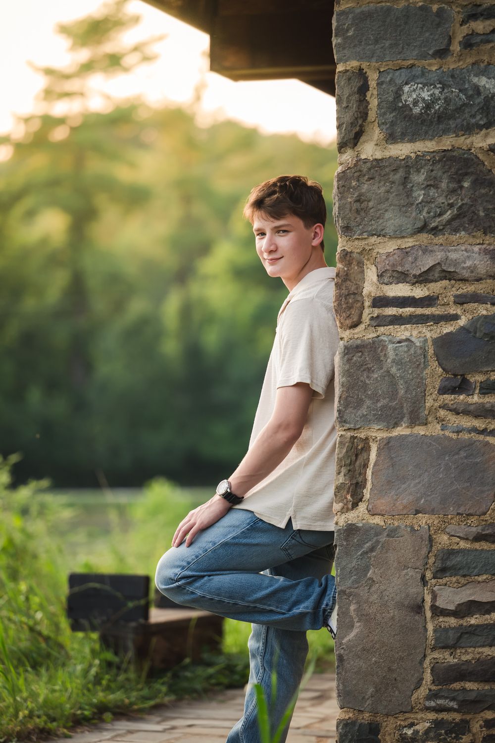 Teen boy in white shirt and jeans leaning against stone pillar outdoors with green trees in background.