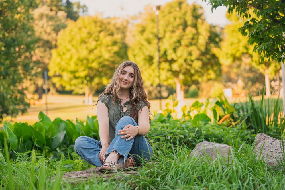 Young woman sitting cross-legged in lush green garden park, smiling, wearing jeans and vest top on sunny day.