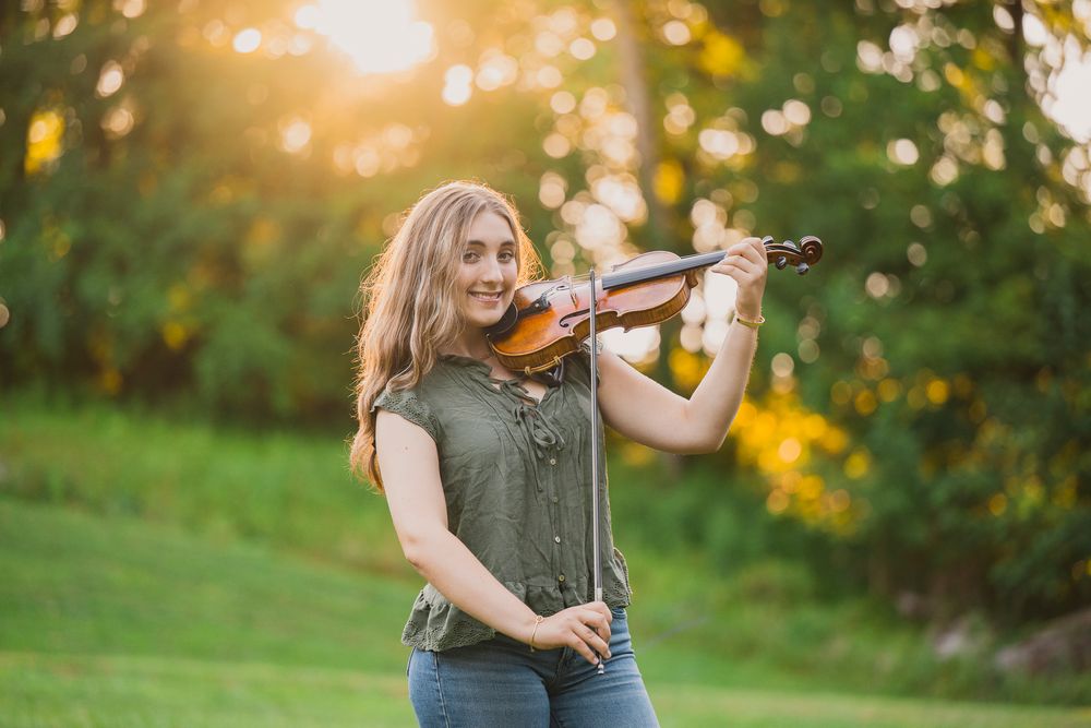 Young woman smiling while playing violin outdoors in golden sunset light, wearing green top and jeans.
