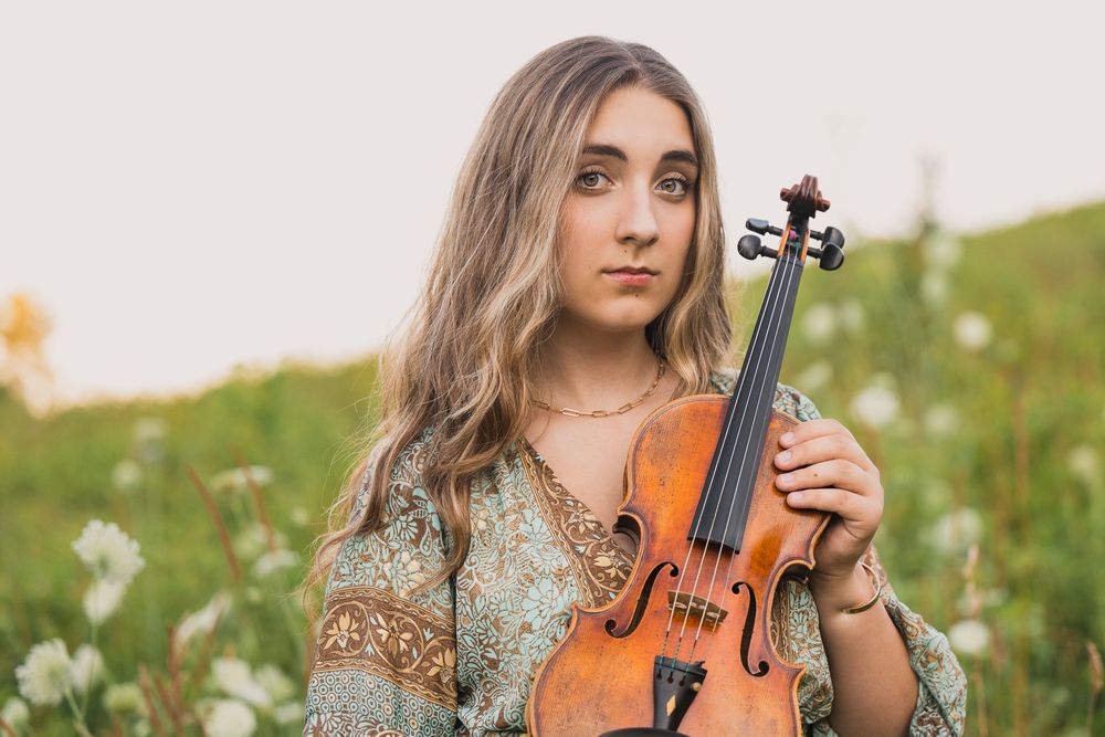 Young woman with long hair holding a violin in a flower field, wearing a boho dress in soft natural light.