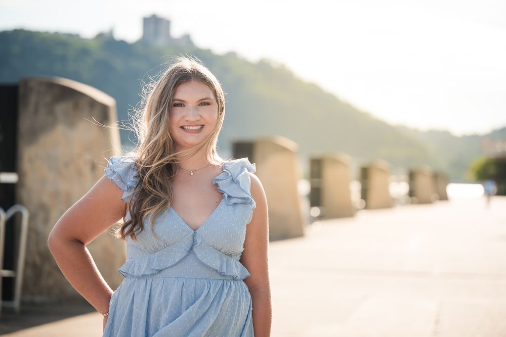 Young woman in light blue ruffled dress smiling outdoors with scenic hillside castle backdrop in warm sunlight.