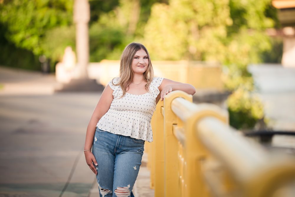 Young woman in ripped jeans and white top leaning on a yellow railing outdoors in warm sunlight.