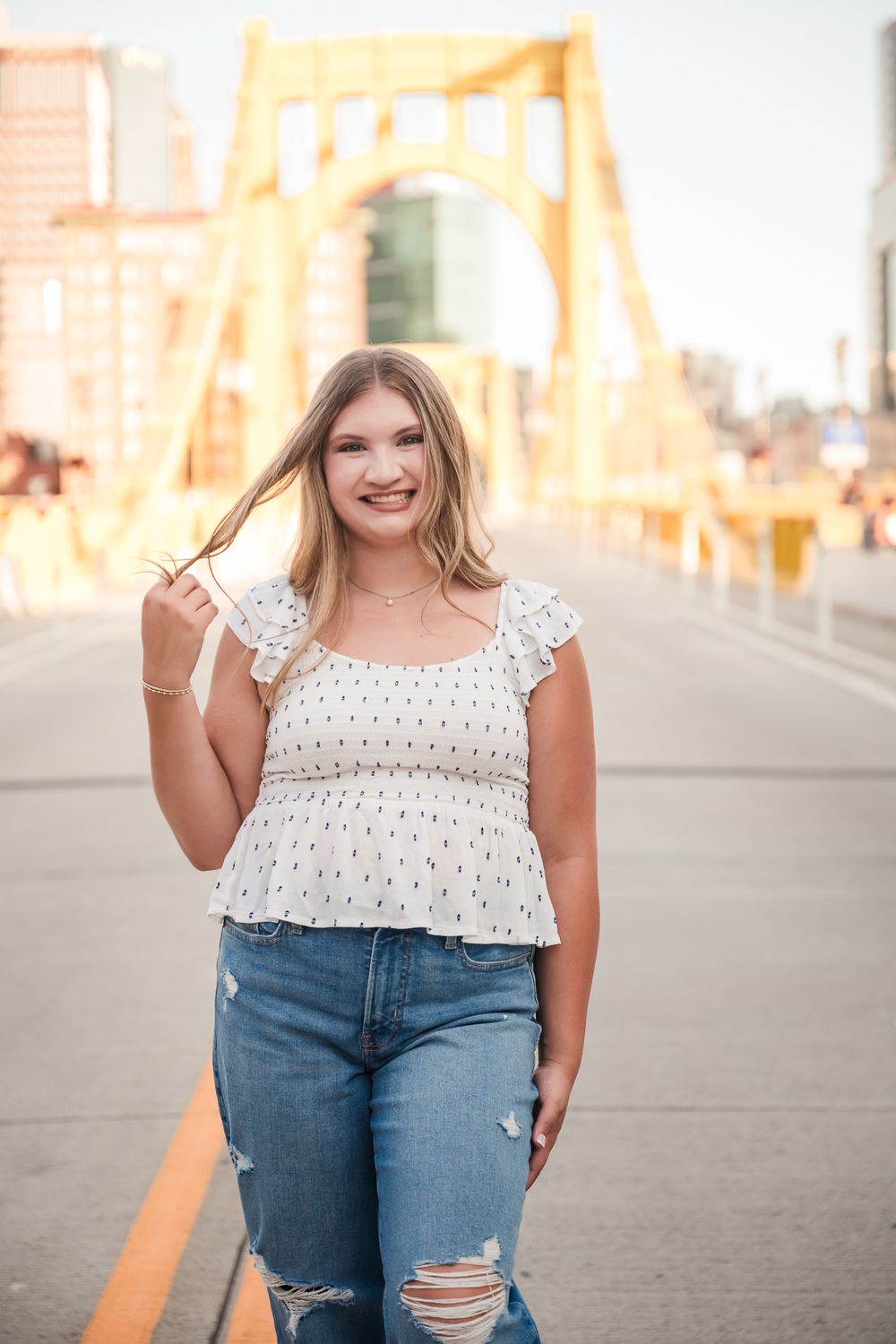 Young blonde woman in white peplum top and ripped jeans smiling on a bridge with yellow arch in Pittsburgh.