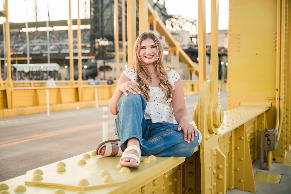 Young blonde woman sitting on a yellow bridge railing, wearing jeans and a white top, smiling outdoors.