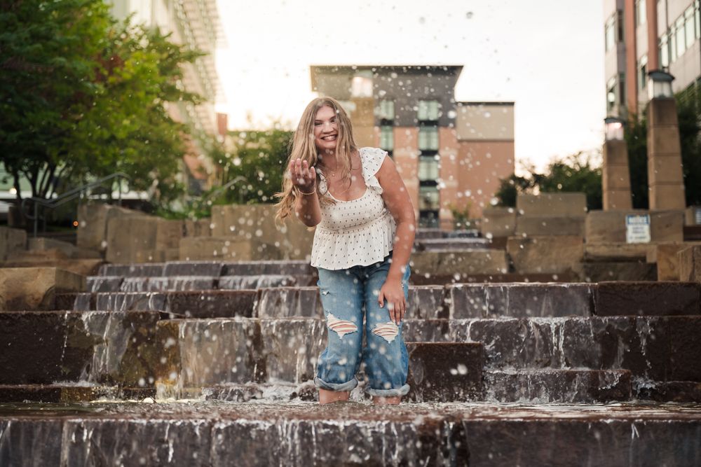 Young woman in ripped jeans and white top posing on urban fountain steps with water splashing around her.