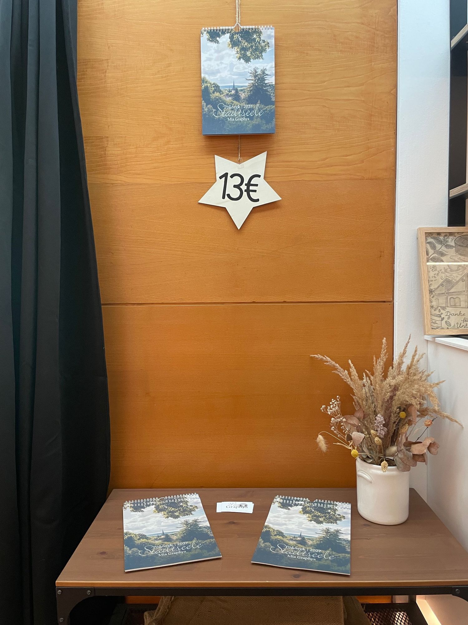 Small wooden shelf display with books priced at 13€, dried flowers in vase, against orange wall with dark curtain.