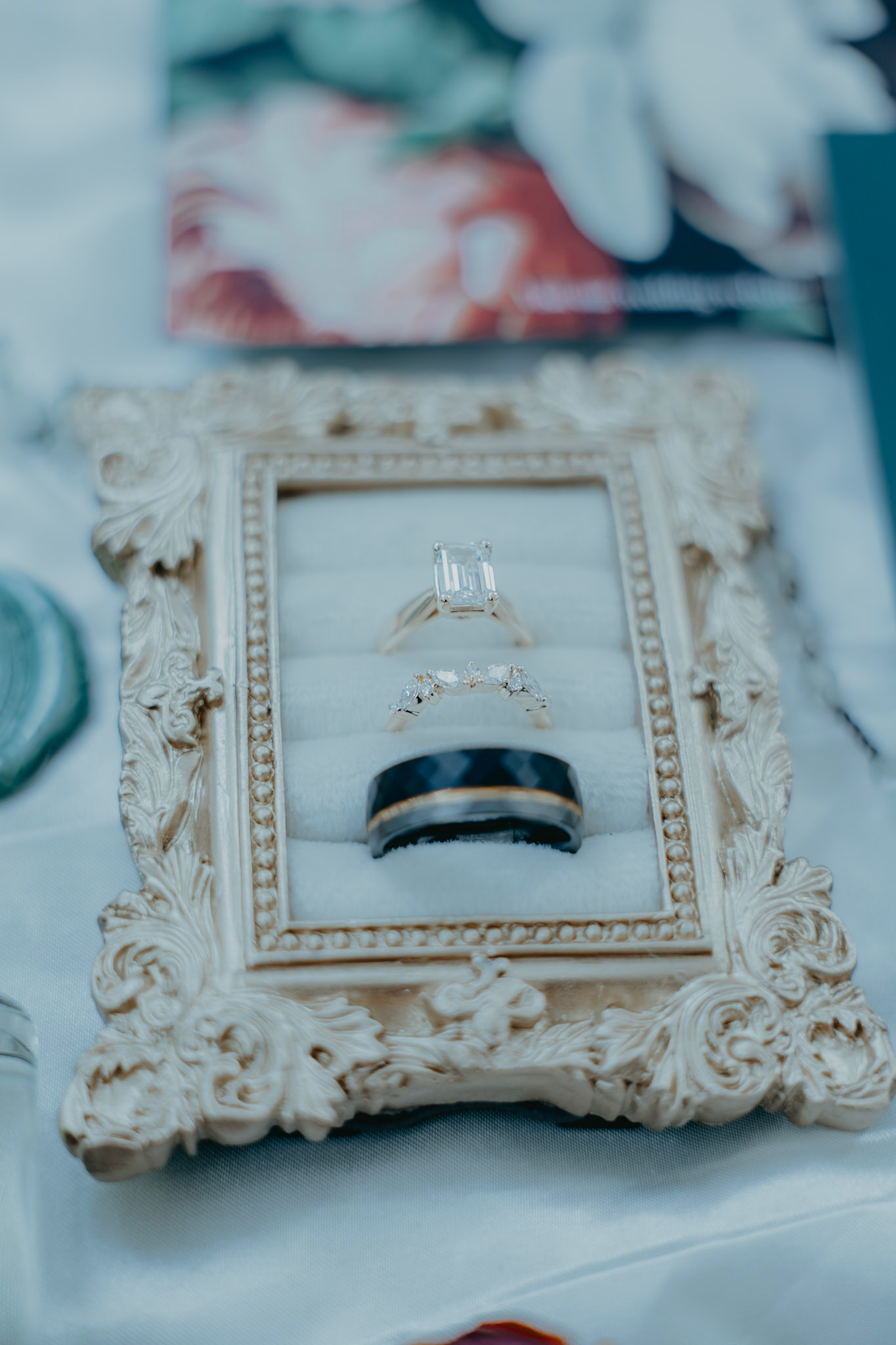 Three wedding rings displayed on an ornate white vintage frame, featuring a diamond engagement ring and two wedding bands.