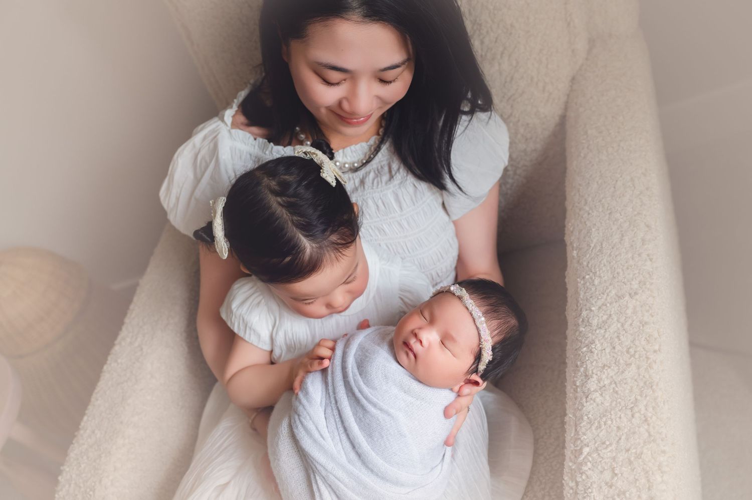 A mother in white dress holds newborn baby and toddler daughter on a cream chair in a soft newborn photo session.