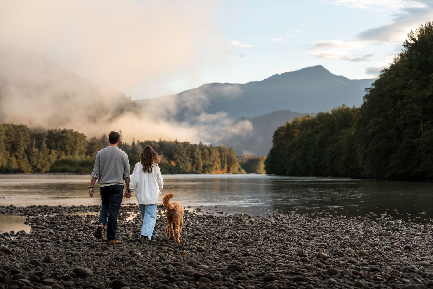 Couple walking with golden dog along rocky riverbank with misty mountains and lush forest in background.