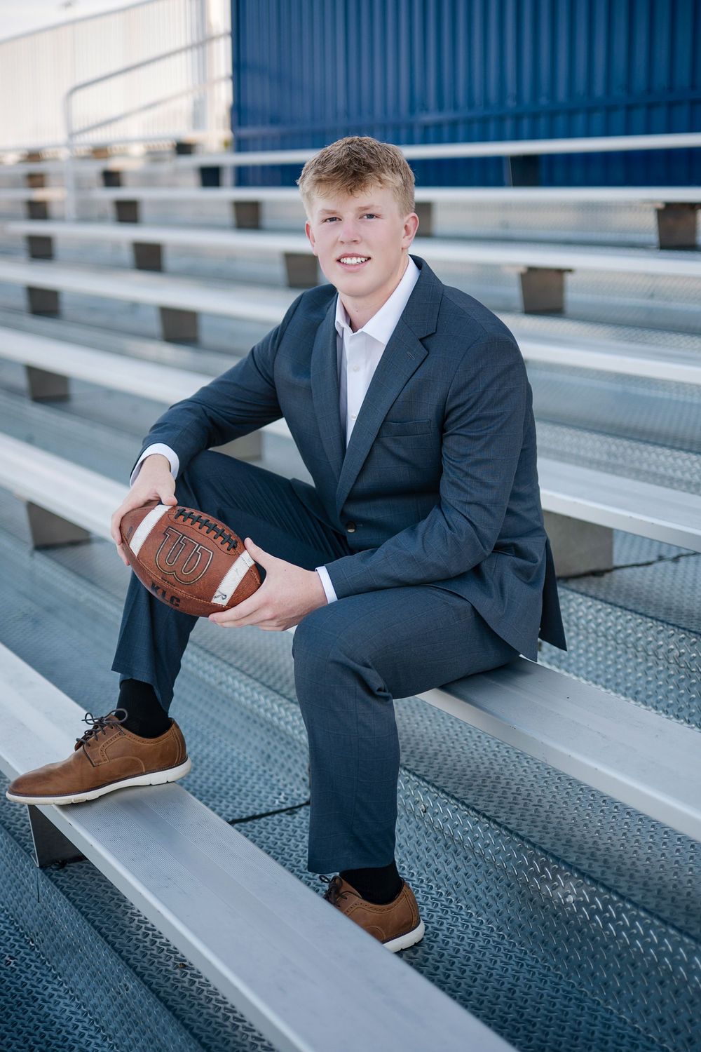 Young man in navy suit holding football while sitting on bleachers, senior portrait photo.