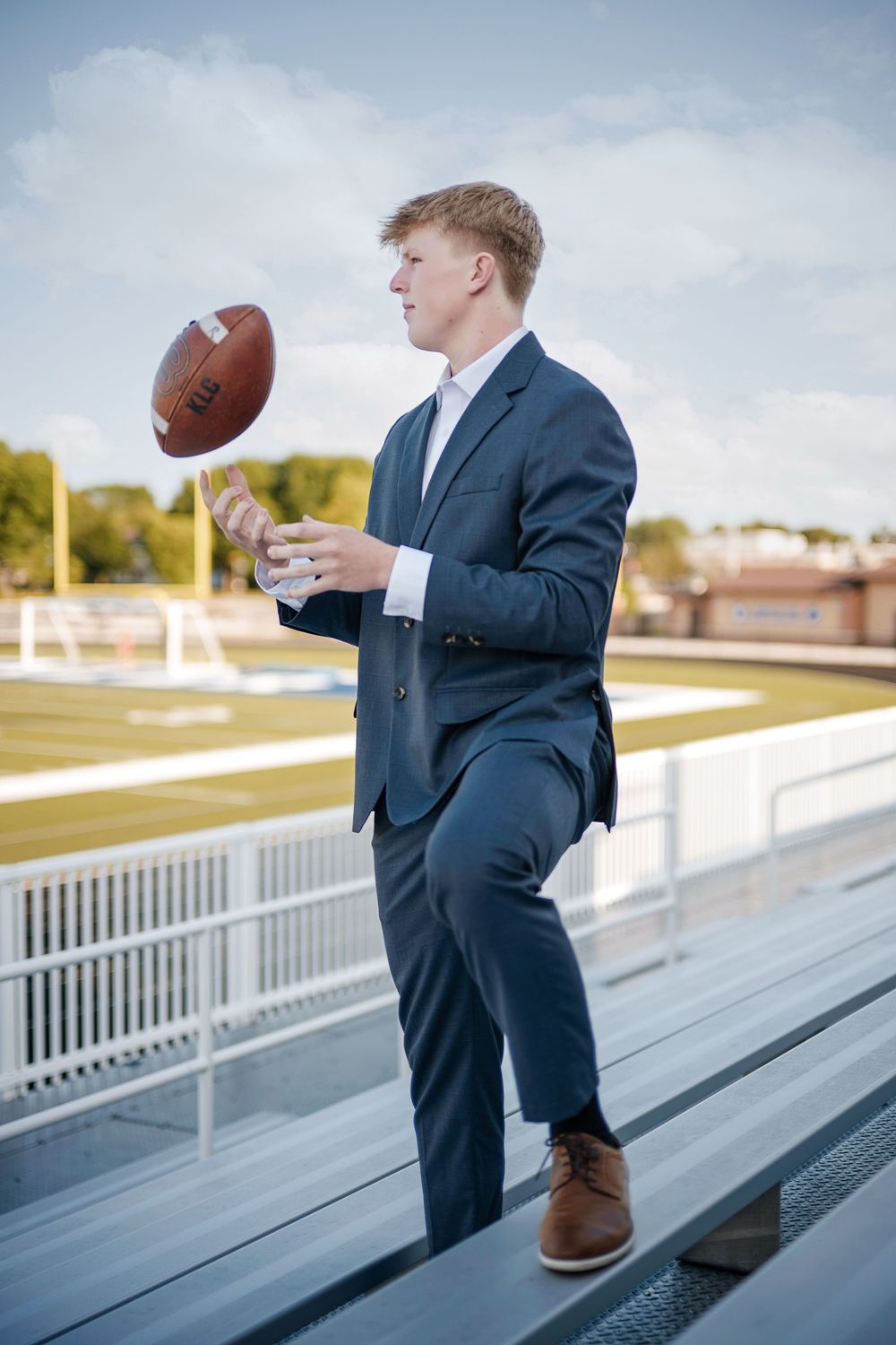 Young man in navy suit tossing a football in stadium bleachers on a sunny day.
