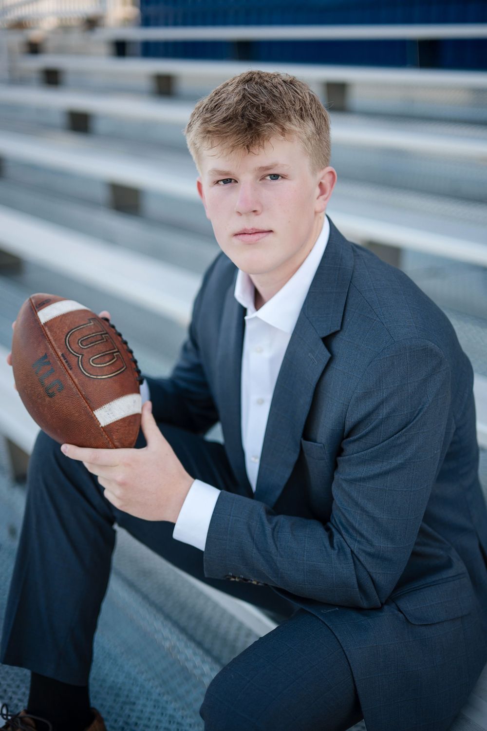 Young man in gray suit holding a Wilson football while sitting on bleachers in a senior portrait photo.