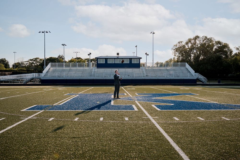 Person standing alone on a blue turf football field with bleachers and stadium lights in background.