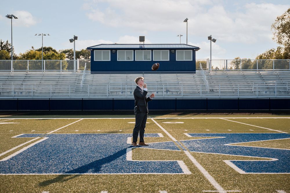 Man in suit standing on blue turf football field with empty bleachers and press box in background.