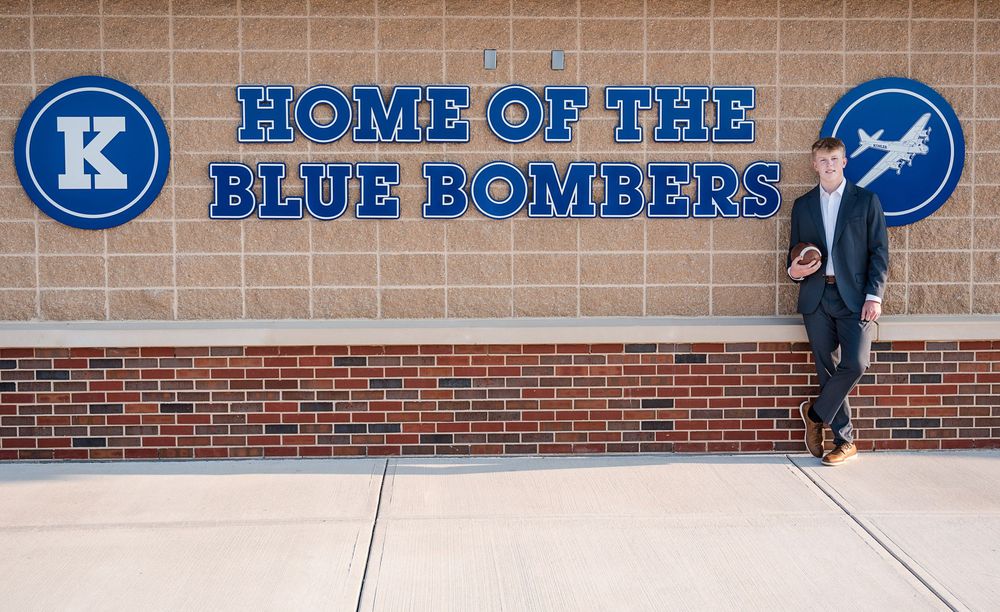 Young man in suit poses by brick wall with Home of the Blue Bombers sign at Kearney school.
